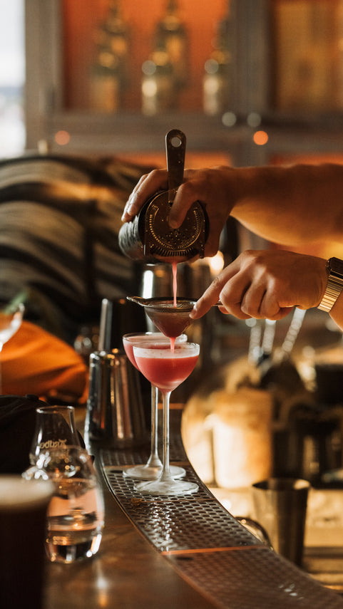 Person pouring a cocktail into a glass at a bar.